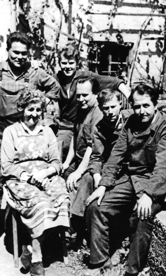 Historic black and white photo of an elderly woman and five men in work clothes in front of a half-timbered house.