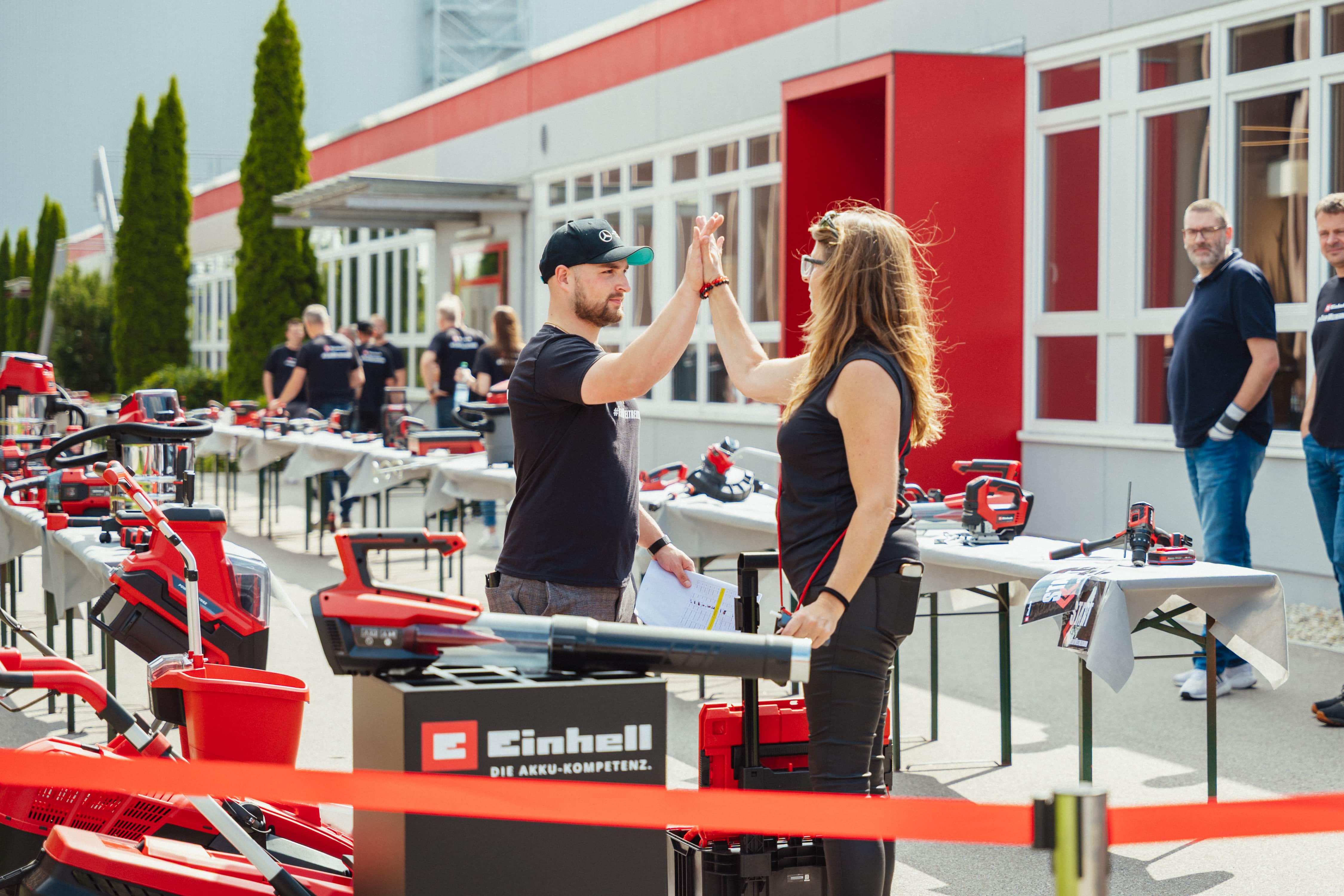 Un hombre y una mujer chocan las manos en un evento al aire libre de Einhell con exposición de herramientas, fotografiado en color.