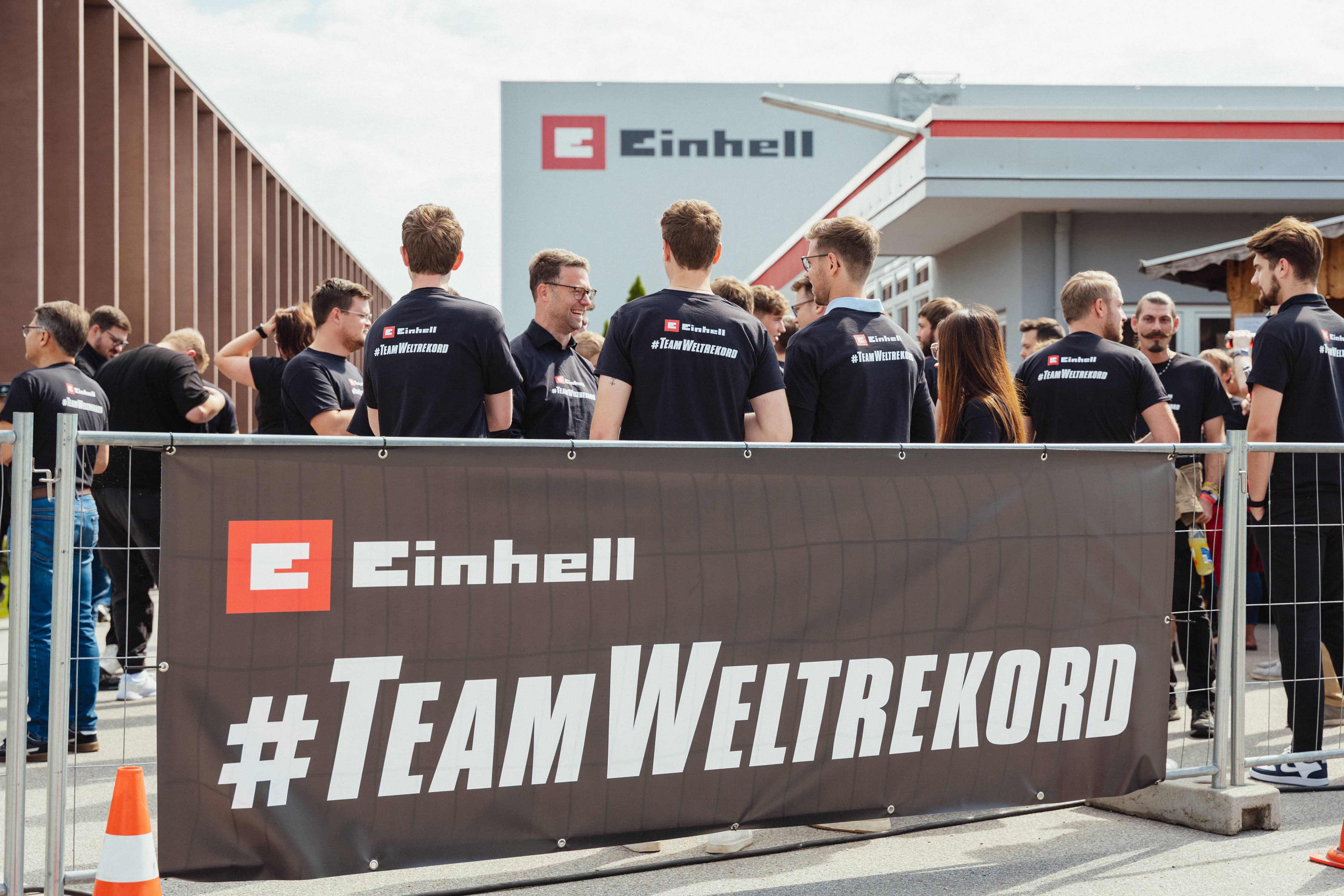 Los participantes se encuentran detrás de una valla con una gran pancarta "Einhell #TeamWorldRecord" en las instalaciones de la fábrica. 