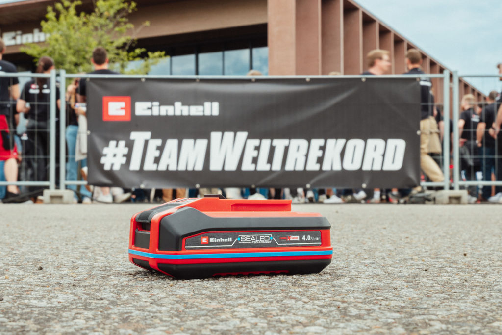 Una batería Einhell SEALED Power X-Change de 4.0 Ah yace sobre el asfalto, con una pancarta "#TeamWorldRecord" al fondo.